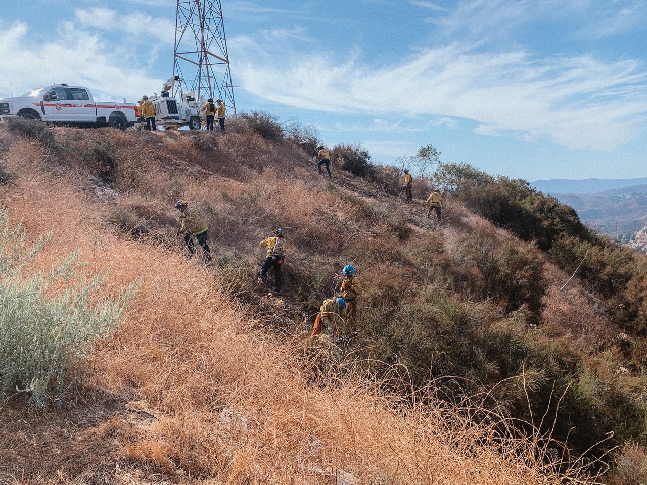A crew working brush along a ridge beneath a transmission corridor, engine staged on the access road above